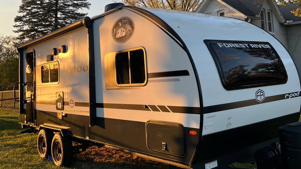 Travel trailer parked on a lawn in low autumn sun, evergreens and fall trees behind it